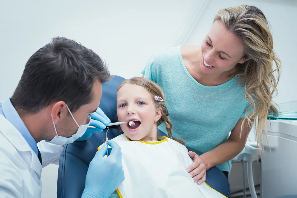 A dentist checking a young girl's teeth while her mother looks on